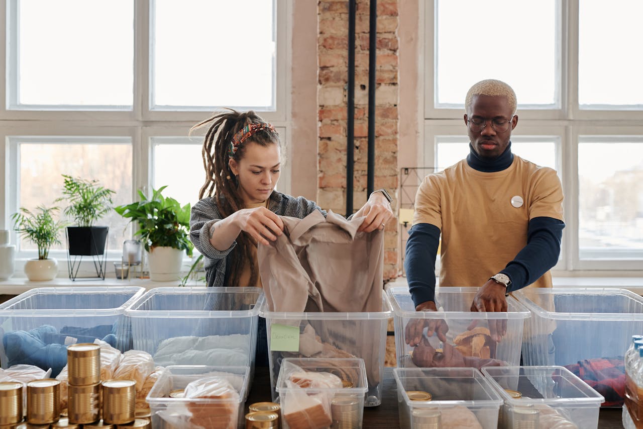 Two volunteers organize clothing and food donations indoors during daylight.